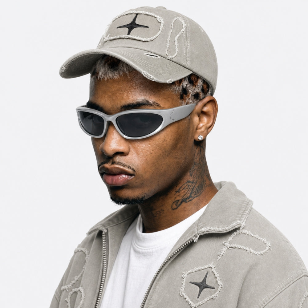A man wearing a light gray distressed baseball cap, matching gray jacket, white t-shirt, and gray cat-eye sunglasses, shown in a close-up profile against a light gray background.