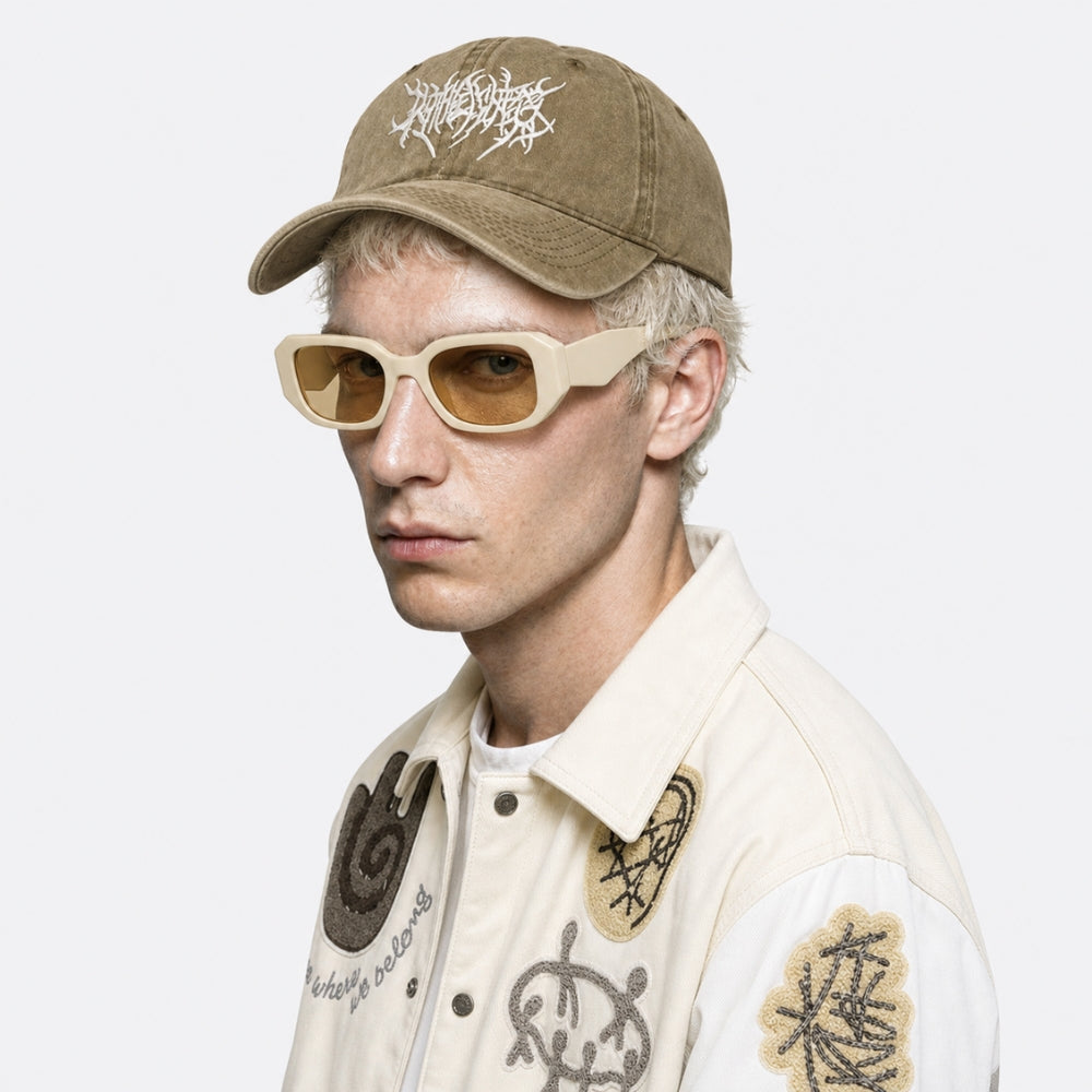 A model wearing a brown baseball cap with a white graphic logo, beige cat-eye sunglasses, and a cream-colored embroidered jacket, shown against a plain light gray background.