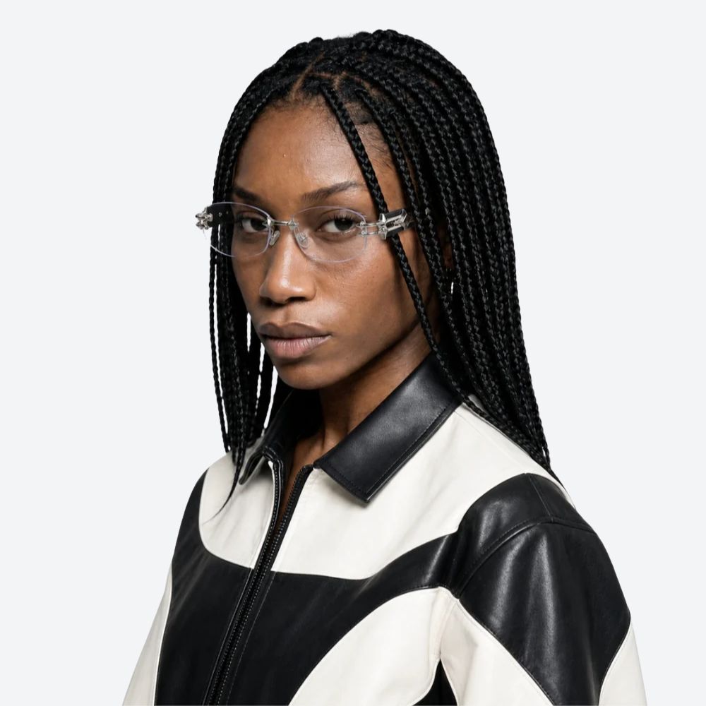 A model with long black box braids and clear-framed eyeglasses, wearing a black and off-white color-blocked jacket with faux leather panels, set against a plain light gray background.