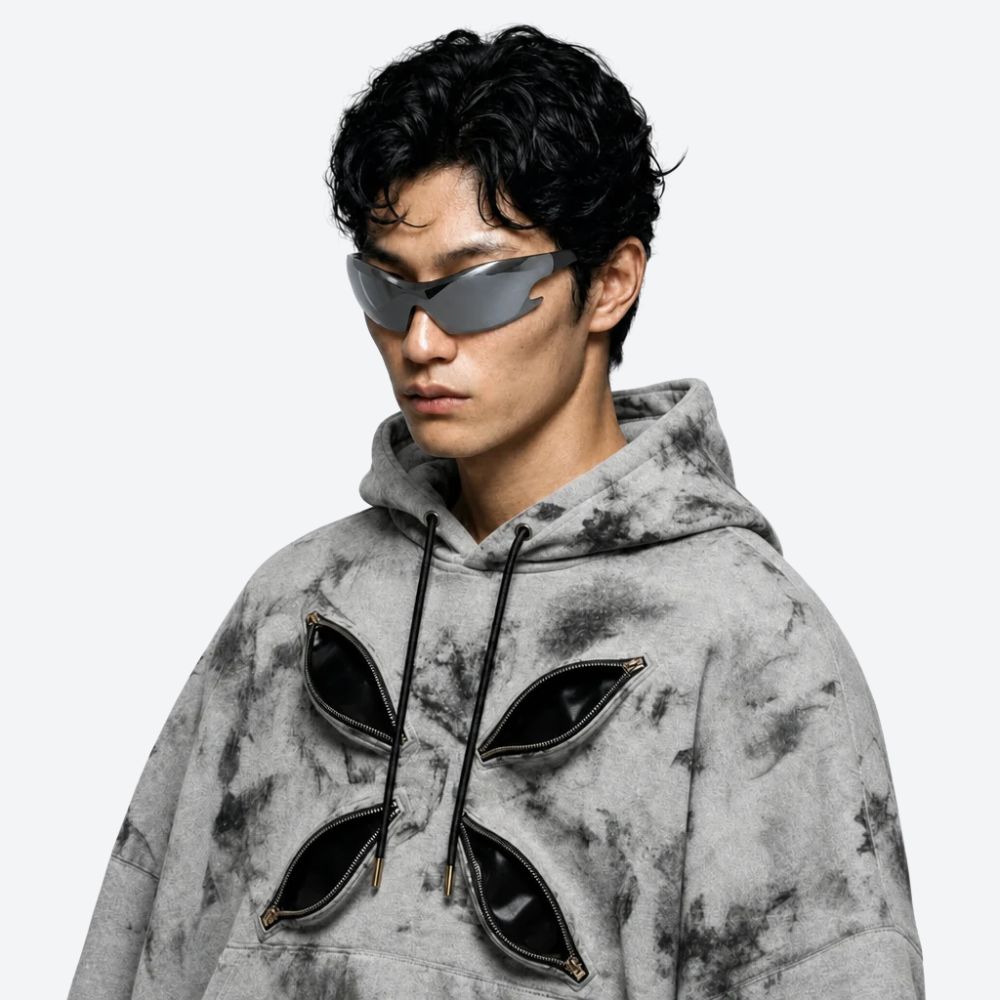 A man wearing a gray and black acid-wash hoodie with four large, stylized black pouches on the front, styled with futuristic street wear gray sunglasses, against a plain light gray background.