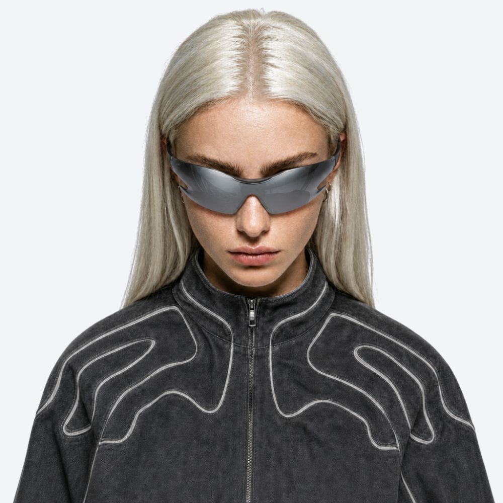 A model with platinum blonde hair wearing futuristic dark gray street wear visor-style sunglasses and a dark gray zip-up jacket with white stitched patterns, set against a solid light gray background.