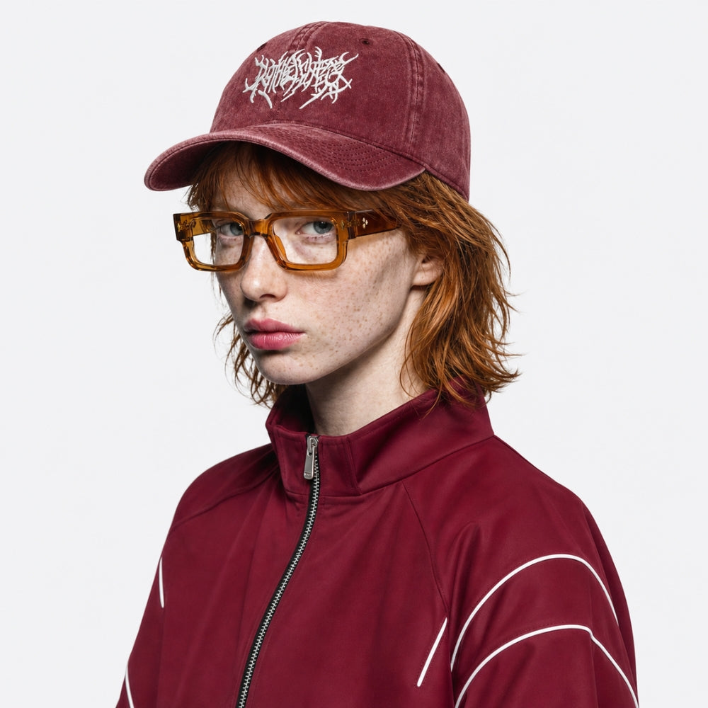 A person wearing a burgundy baseball cap with a white stylized logo, amber-framed glasses, and a matching burgundy zip-up jacket with white piping, photographed against a light gray studio background.