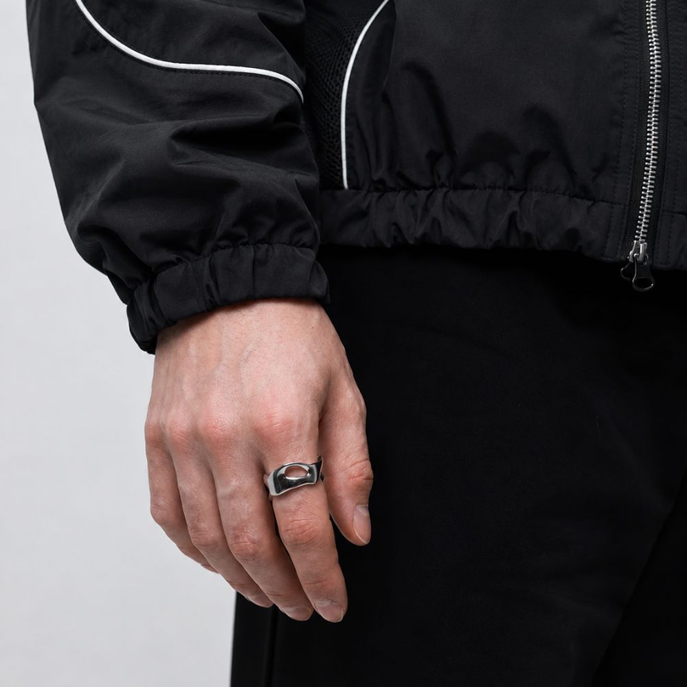 A close-up of a person's hand wearing a silver-toned metal ring with a black and red geometric design, styled with a black windbreaker jacket and black pants.