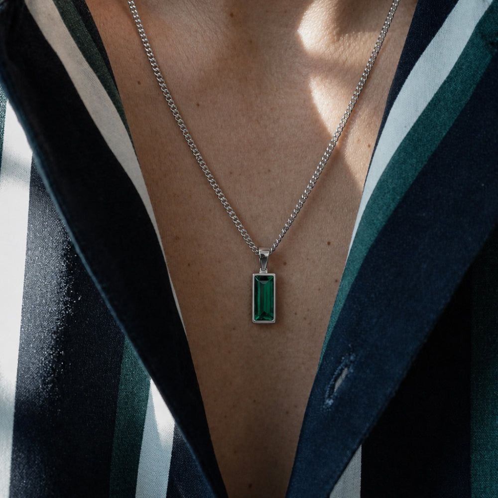 A close-up of a person wearing a silver chain necklace with a rectangular green emerald-cut pendant, styled with a dark striped blazer.
