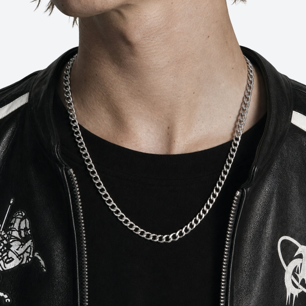 A close-up of a person's neck and upper chest wearing a thick silver curb chain necklace over a black t-shirt and black leather jacket.