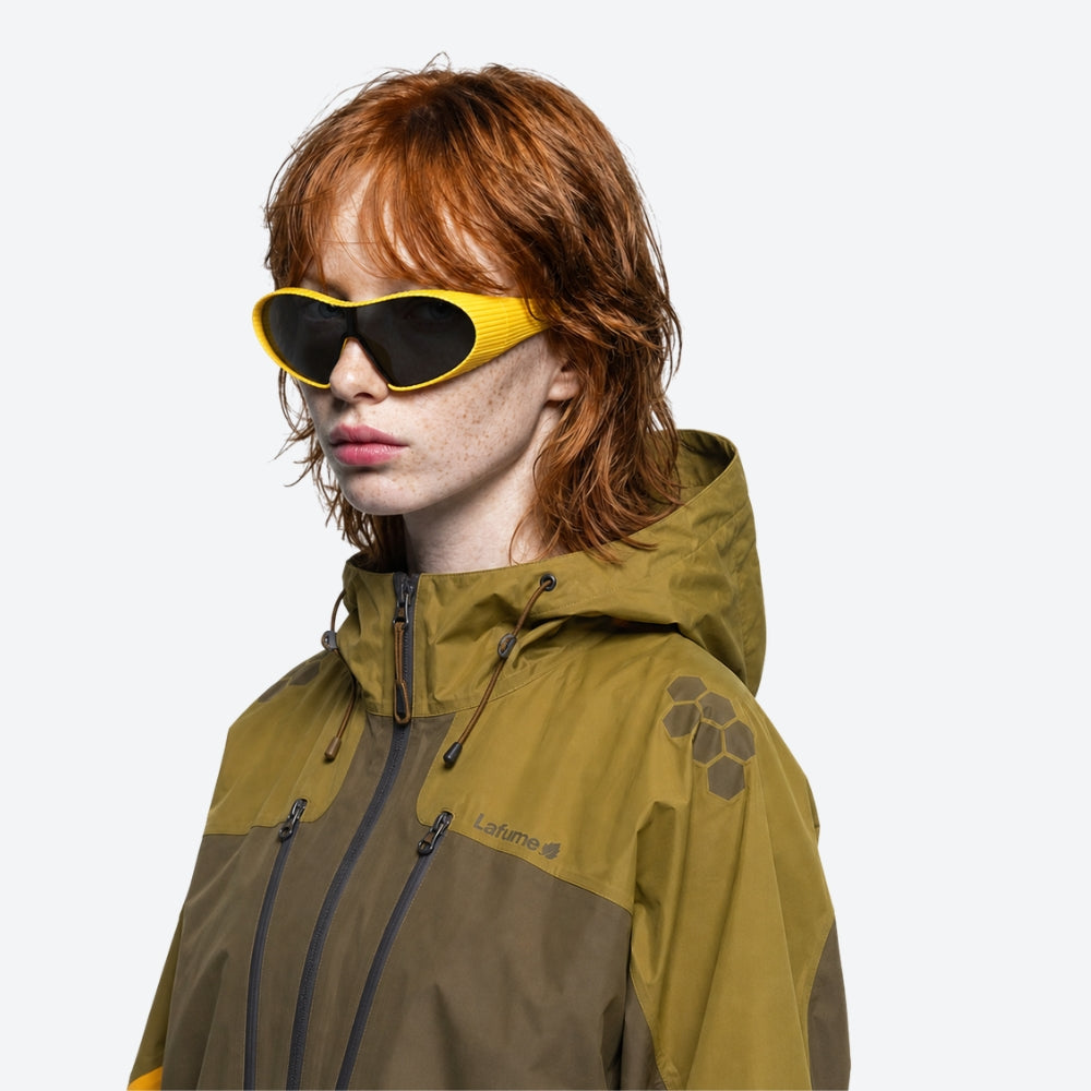 Person wearing a green and brown jacket with a brand logo, wearing yellow VYSER Yellow sunglasses with black lenses on a light gray background