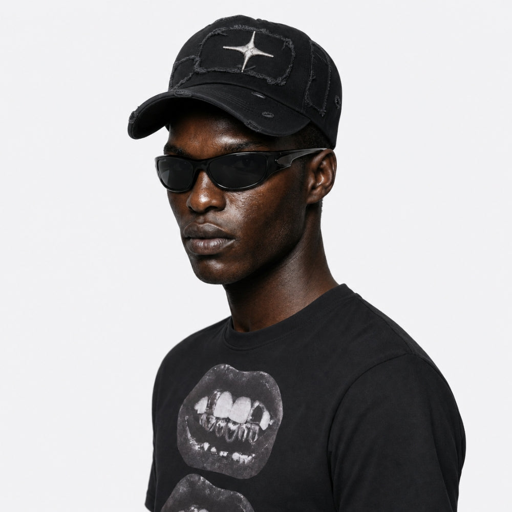 A man wearing a black baseball cap with a distressed star logo, black sunglasses, and a black graphic t-shirt, shown in a side profile against a light gray background.