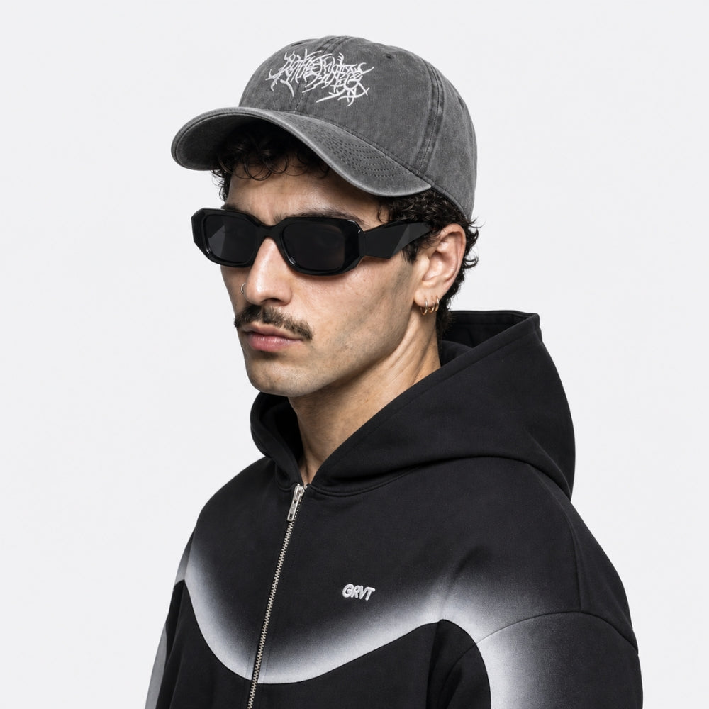 A man wearing a dark gray baseball cap with a white graphic logo, black sunglasses, and a black gradient zip-up hoodie, shown in a side profile against a light gray background.