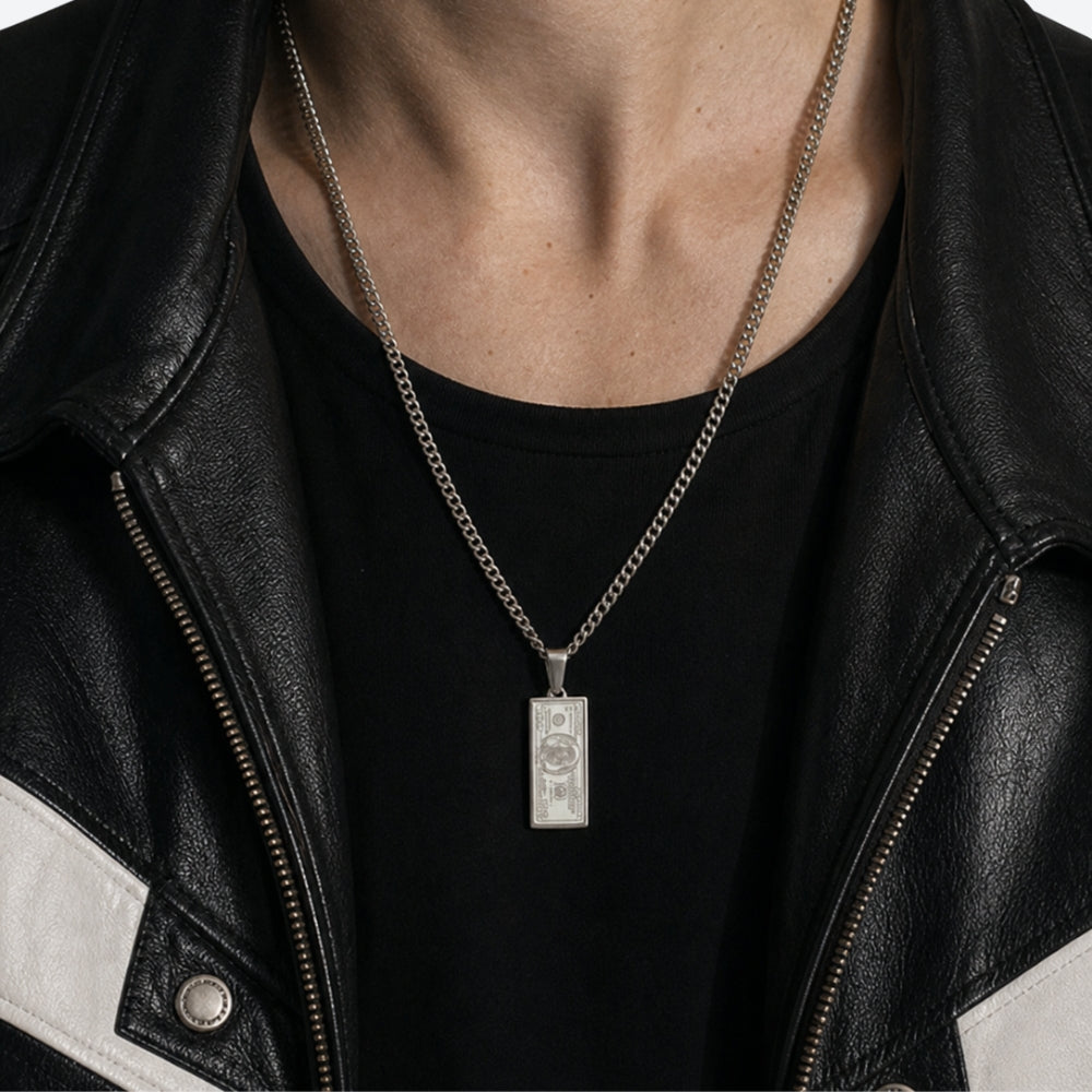 A silver-toned rectangular pendant necklace with an engraved dollar-bill design, worn on a model's neck over a black t-shirt and black-and-white leather jacket.