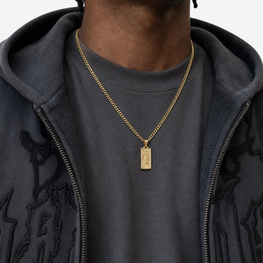 A close-up of a person wearing a gold chain necklace with a rectangular pendant, styled over a dark gray t-shirt and a black embroidered zip-up hoodie.