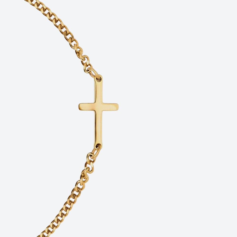 GOLD. A close-up of a gold chain bracelet with a small polished cross pendant, light gray background.
