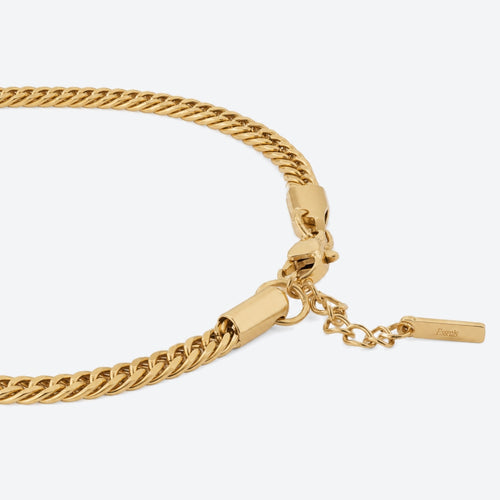 Close-up of a gold-toned chain necklace with a lobster clasp, extension chain, and a small rectangular brand tag on a light gray background.