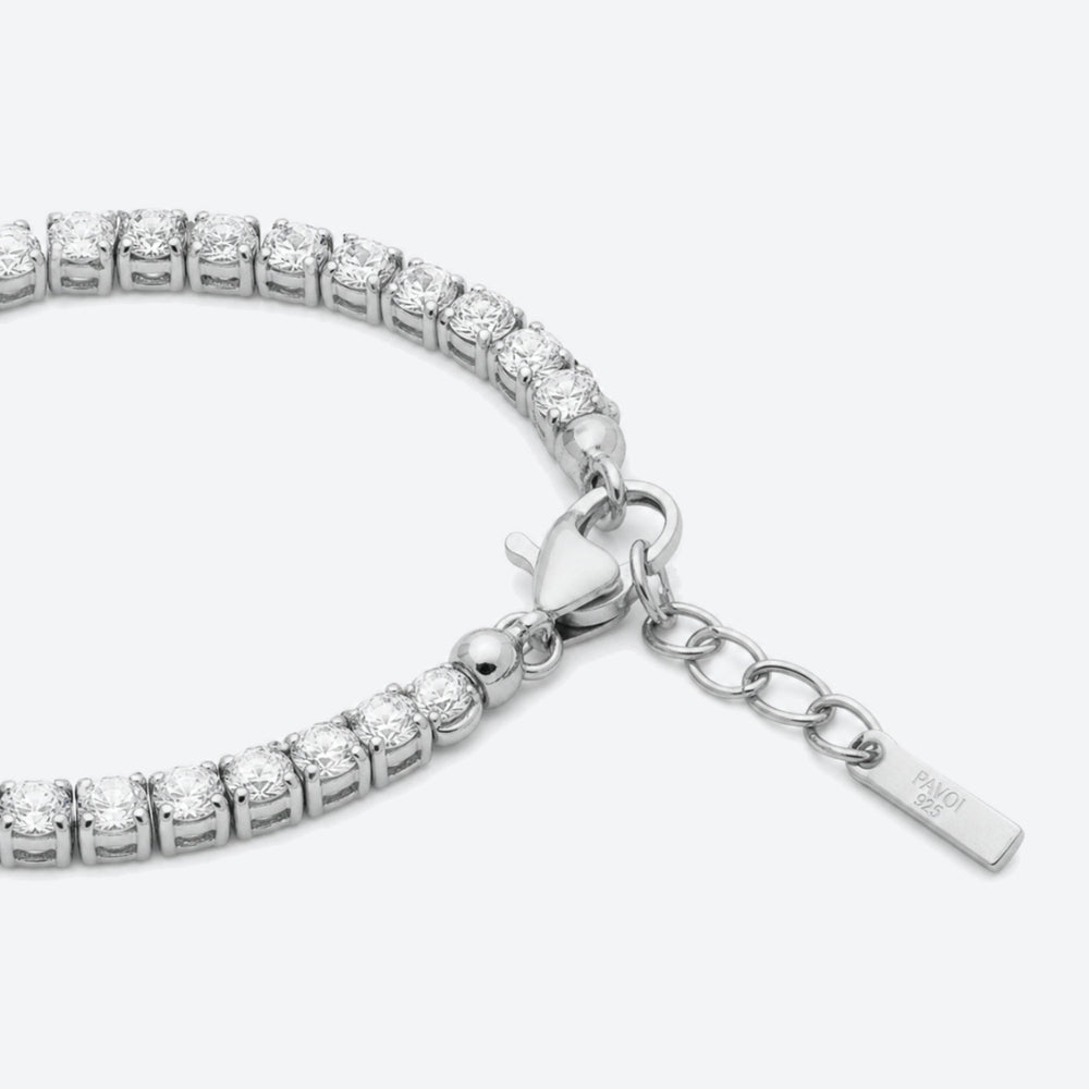 A close-up of a tennis bracelet made of silver-toned metal and clear stones, showing a heart-shaped clasp, extension chain, and a small engraved tag on a light gray background.