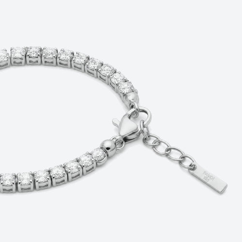 A close-up of a tennis bracelet made of silver-toned metal and clear stones, showing a heart-shaped clasp, extension chain, and a small engraved tag on a light gray background.