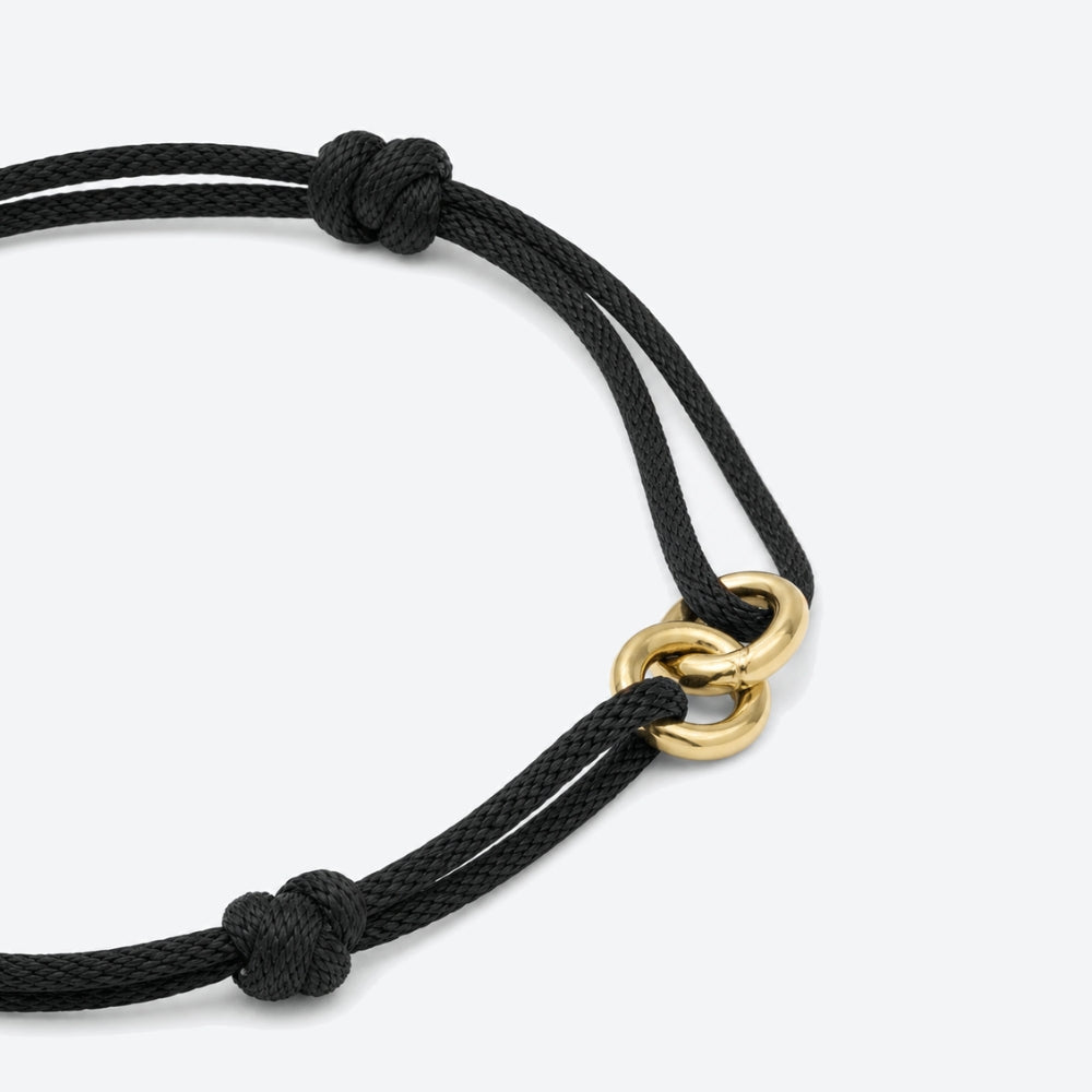 A close-up of a black braided cord bracelet with two knotted ends and a polished gold interlocking ring clasp, set against a clean light gray background.