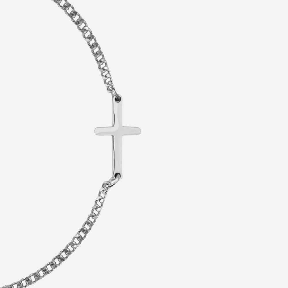 A silver-toned cross pendant on a curb chain bracelet, shown against a light gray background.