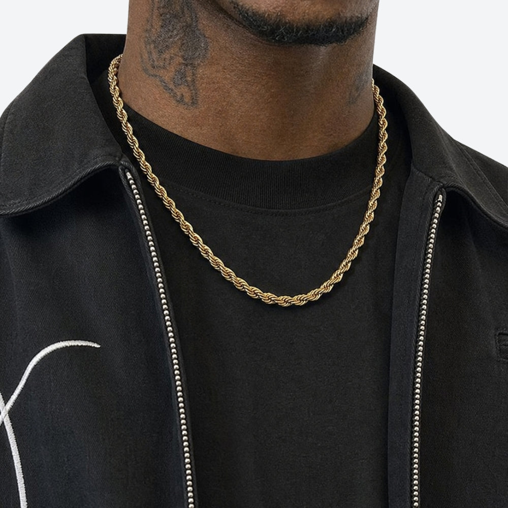 A close-up of a man wearing a gold rope chain necklace over a black t-shirt and black zip-up jacket.