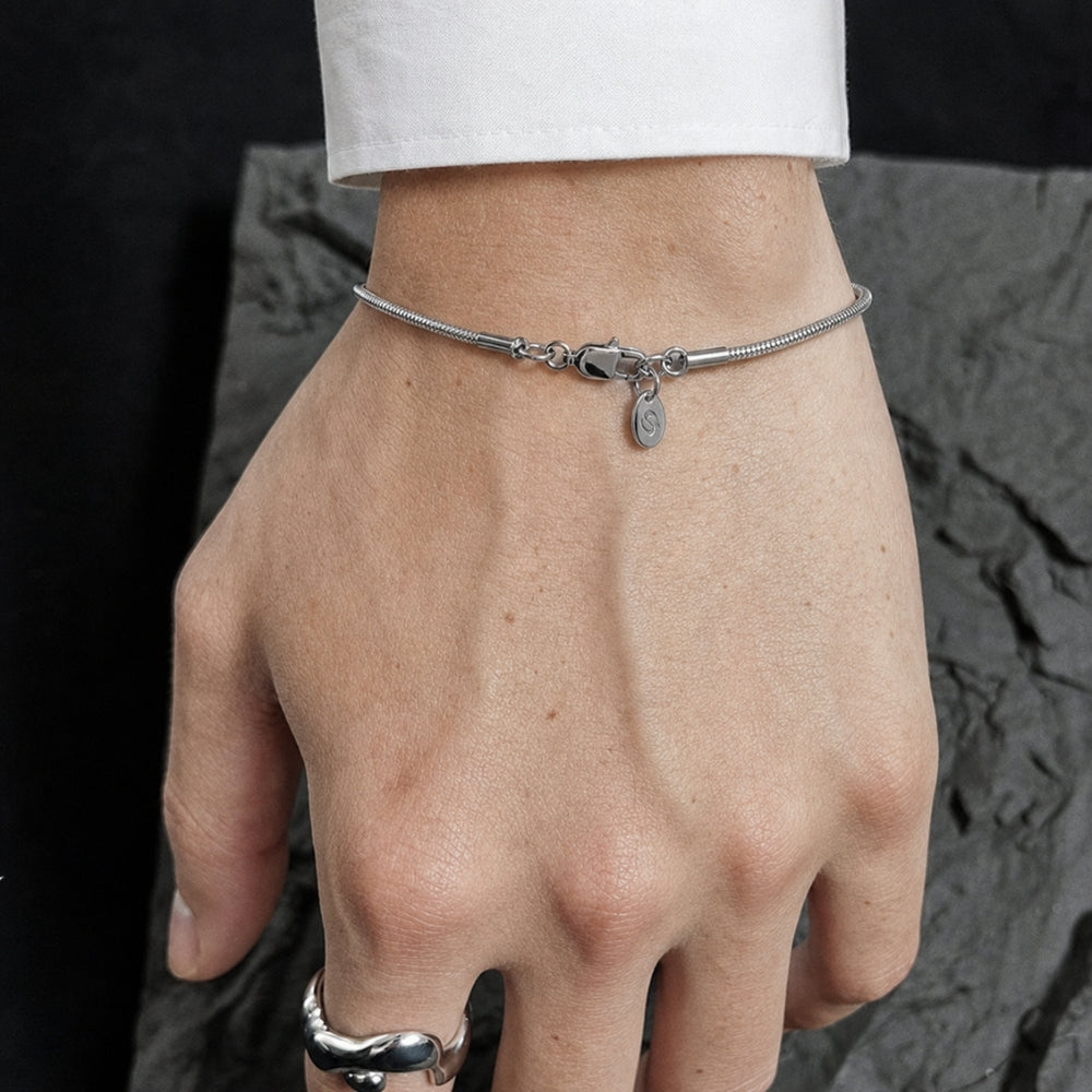 A silver snake chain bracelet with a small oval charm, worn on a person's wrist. The person is wearing a white long-sleeve shirt and a silver ring on one finger, set against a dark textured stone background.