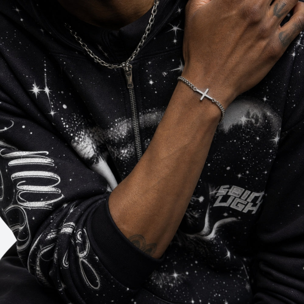 A close-up lifestyle shot of a person wearing a silver chain bracelet with a cross pendant, styled with a matching silver chain necklace and a black zip-up hoodie featuring a white galaxy print.