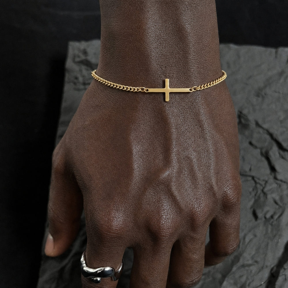 GOLD. A gold cross chain bracelet worn on a wrist resting on dark slate, black background.
