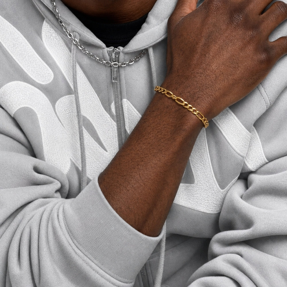 A close-up of a person wearing a gold chain link bracelet on their wrist, paired with a silver chain necklace and a light gray hoodie with a white abstract pattern.