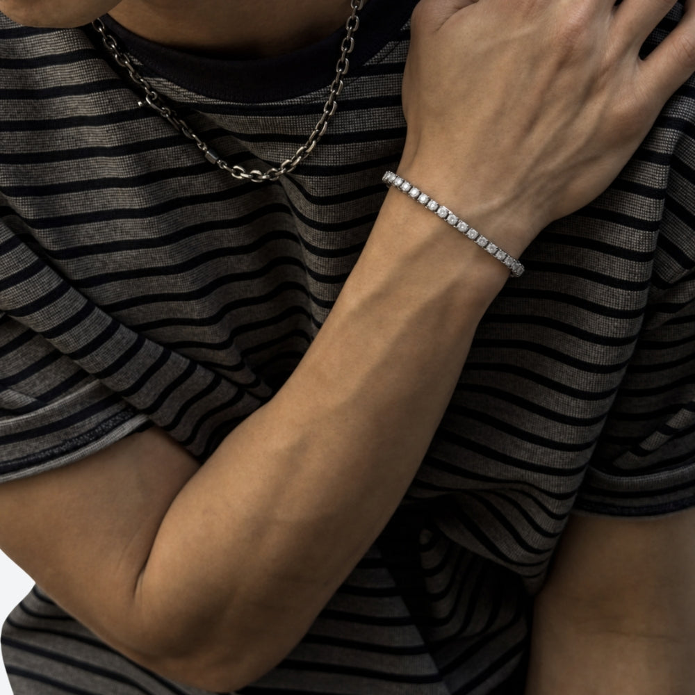 A close-up of a person wearing a delicate silver bracelet with small clear stones, paired with a silver chain necklace and a black-and-gray striped shirt.