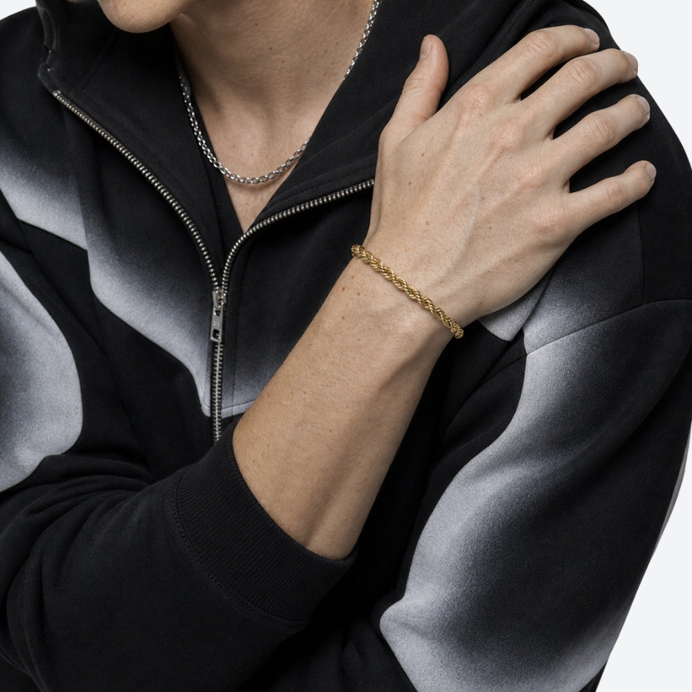 A close-up of a person wearing a gold braided chain bracelet on their wrist, paired with a silver chain necklace and a black and gray gradient zip-up hoodie.