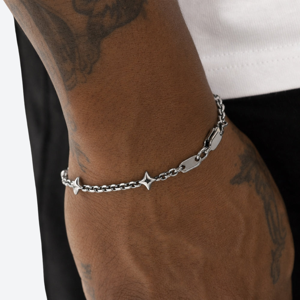 A close-up of a person's tattooed wrist and hand wearing a silver chain bracelet with star and tag charms, paired with a white t-shirt and black pants.