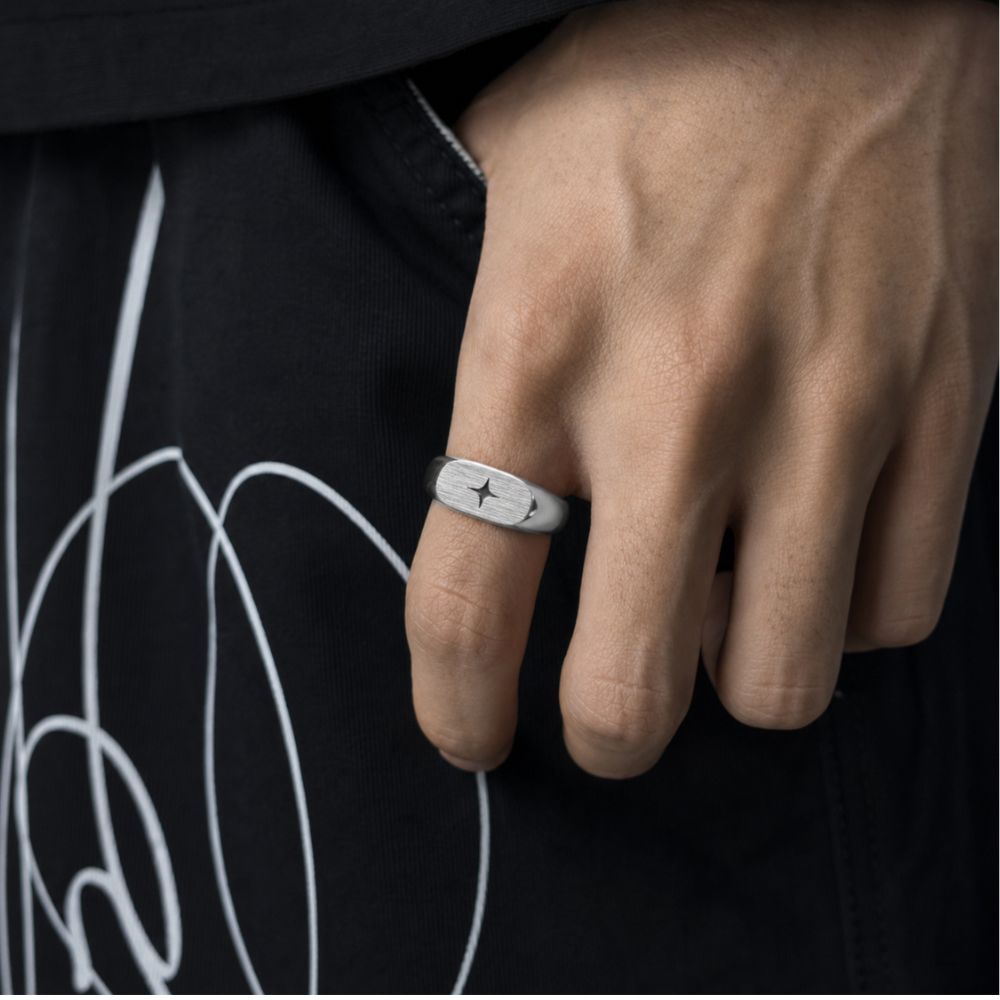 A close-up of a person's hand wearing a silver band ring with a star engraving, set against a black t-shirt with white graphic lines.