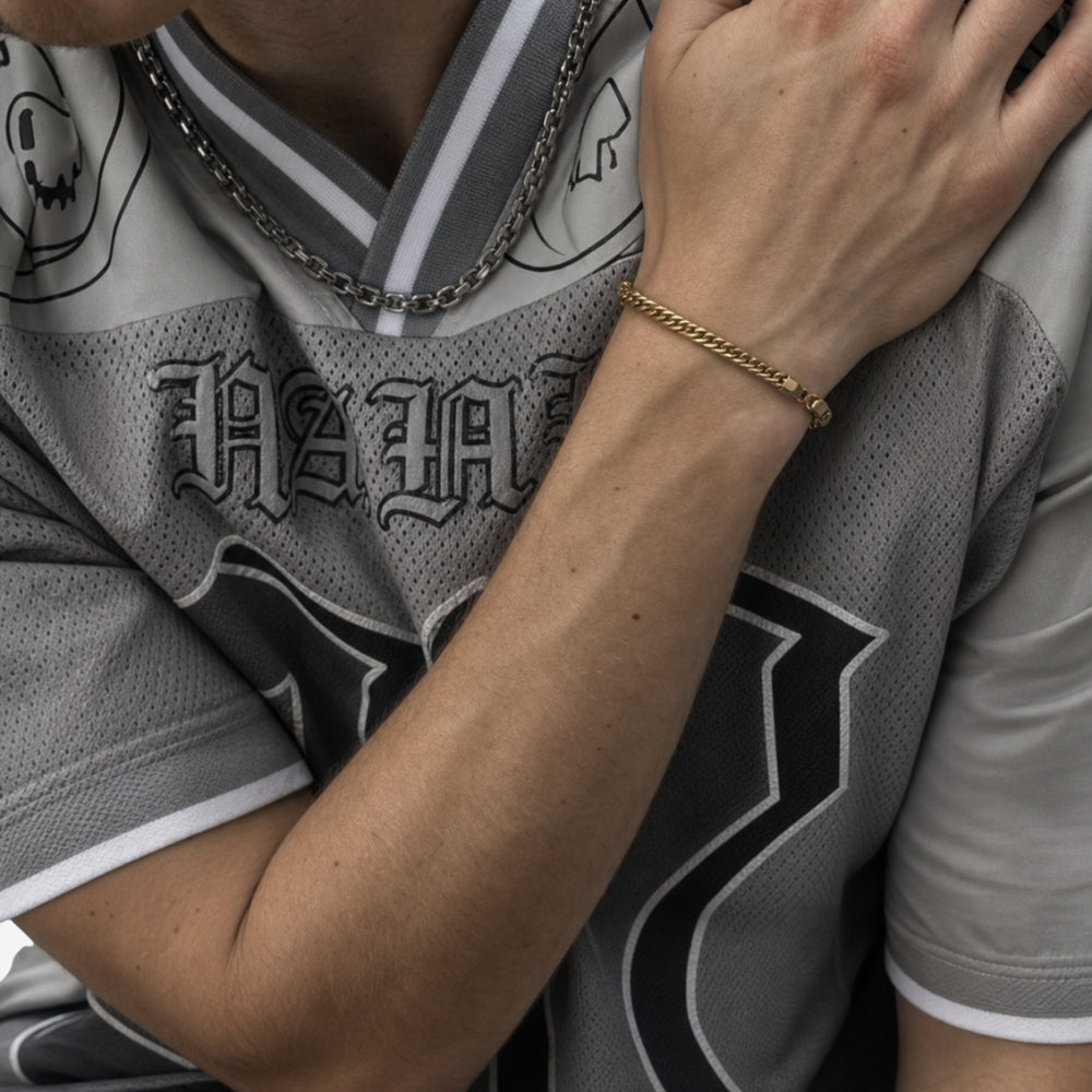 A close-up lifestyle shot of a person wearing a gold chain bracelet on their wrist, a silver chain necklace, and a gray and black sports jersey.