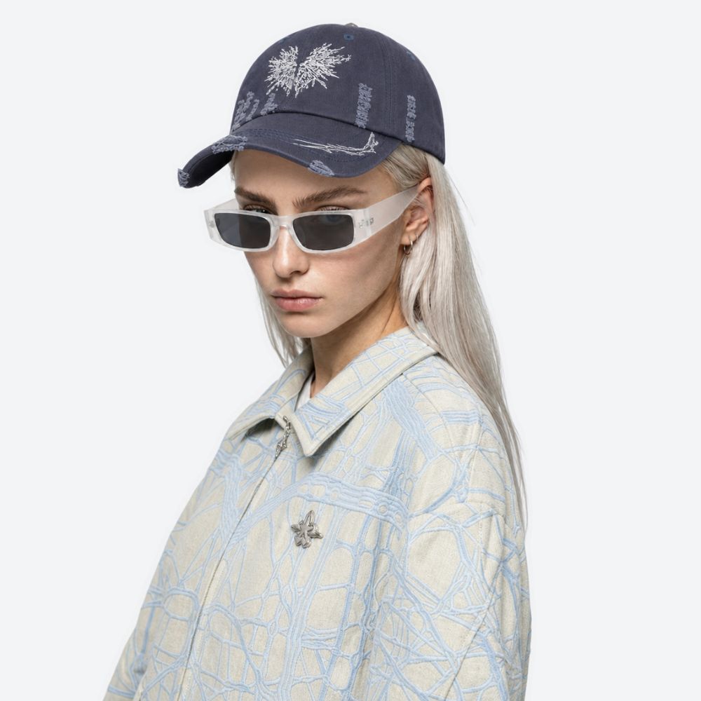 A woman wearing a navy blue moth-embroidered baseball cap with sunglasses and a light patterned jacket