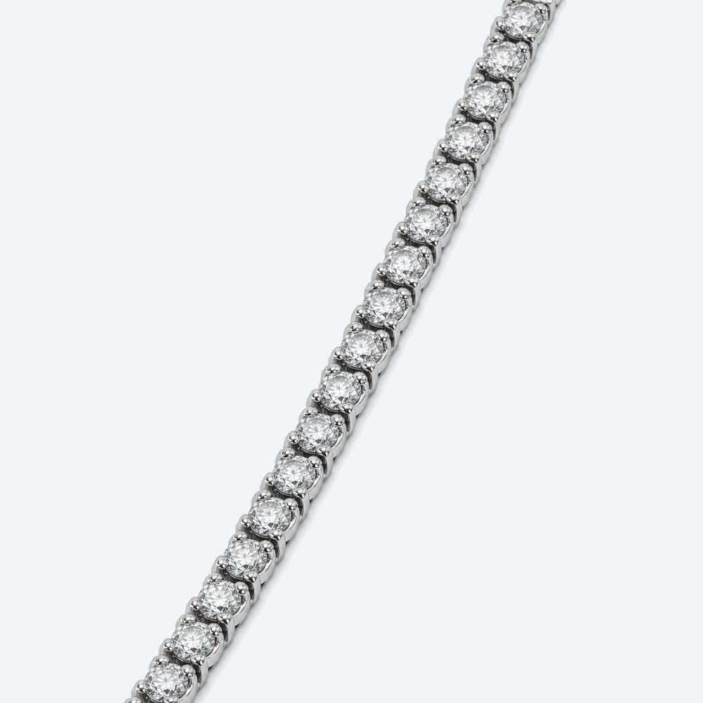 A close-up diagonal view of a silver tennis bracelet with round-cut clear stones, set against a plain light gray background.