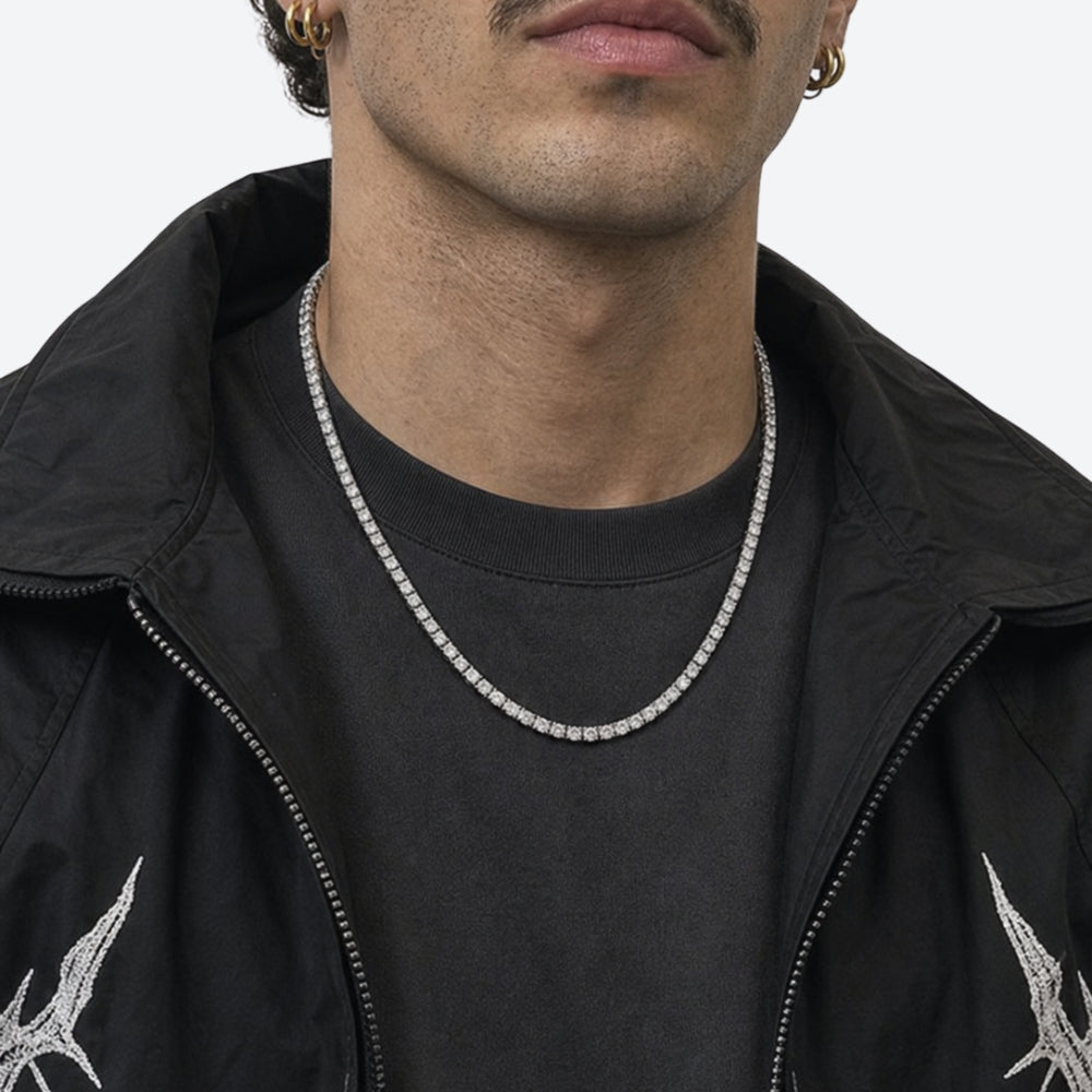 A close-up of a man wearing a silver-toned diamond-look tennis necklace over a black t-shirt and black jacket.