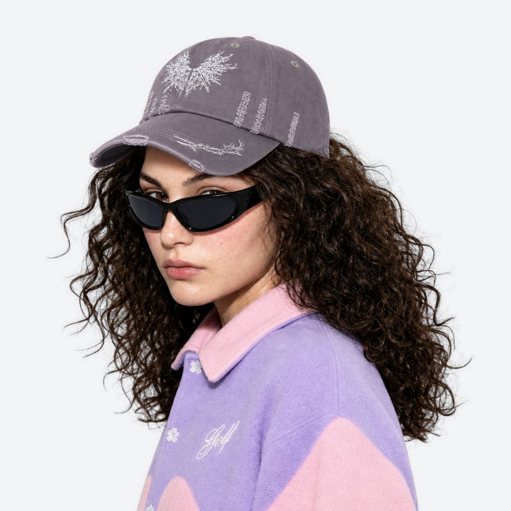 A woman wearing a grey moth-embroidered baseball cap with sunglasses and a lilac top