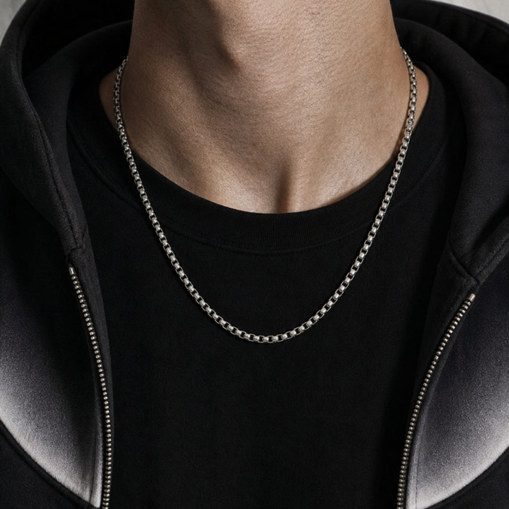 SILVER. A person wearing a silver-toned box chain necklace over a black t-shirt and black zip-up hoodie.