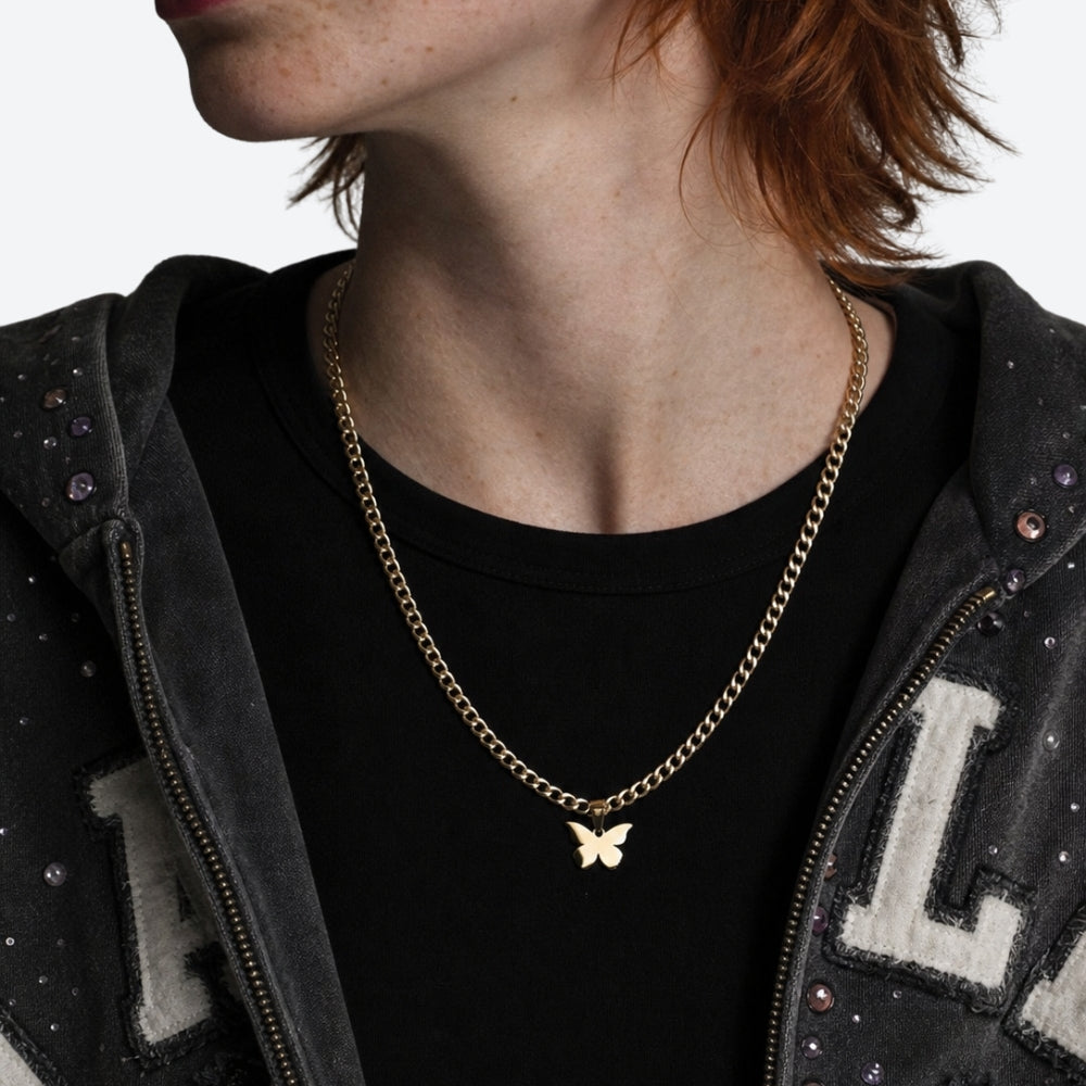 A close-up of a person wearing a delicate gold chain necklace with a small butterfly pendant, styled over a black t-shirt and a dark gray studded zip-up hoodie.