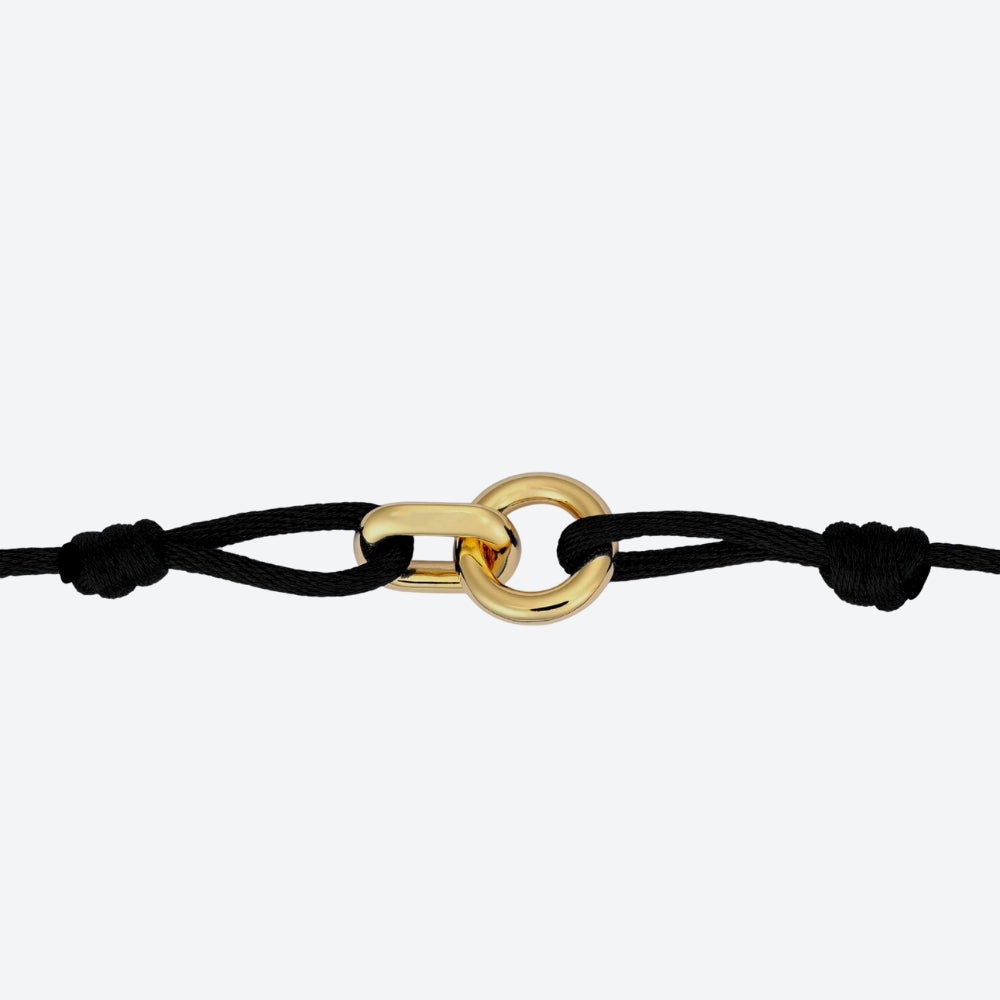 A minimalist black cord bracelet featuring a polished gold interlocking ring and chain-link style clasp, shown against a light gray background.