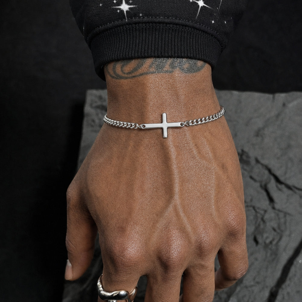 A close-up of a person's hand wearing a silver chain bracelet with a small cross pendant, paired with a silver ring, set against a dark textured background.