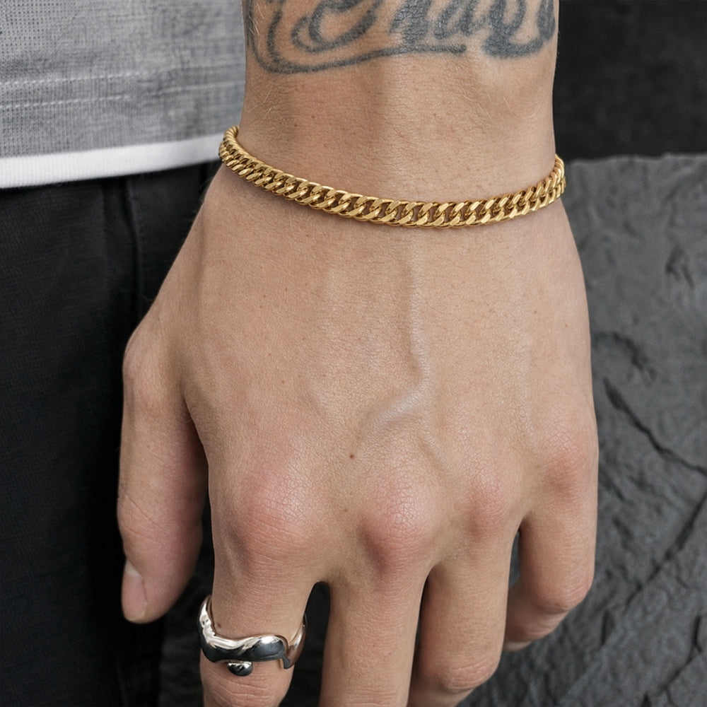 A close-up of a person's hand wearing a thick gold Cuban link chain bracelet, with a silver ring on the finger and a gray shirt and dark textured background visible.