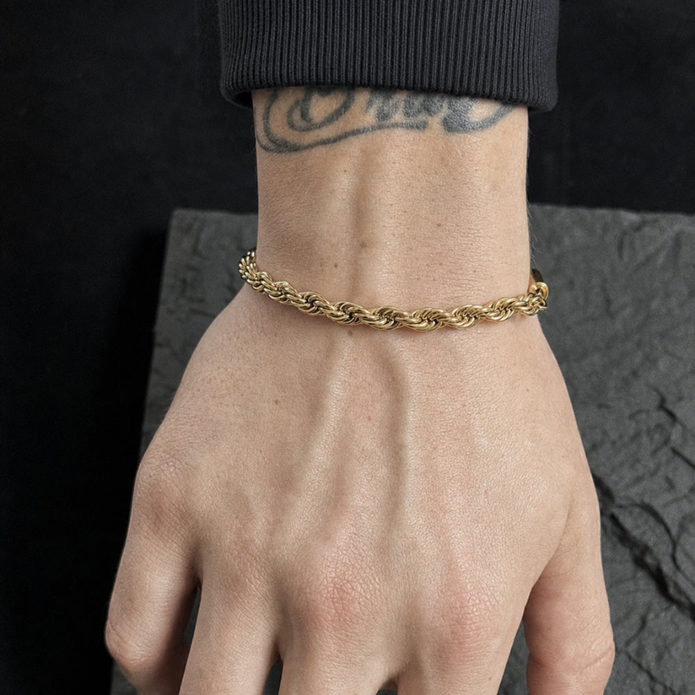A close-up of a person's wrist and hand wearing a thick, twisted gold chain bracelet. The hand has a visible tattoo, and the person is wearing a black ribbed long-sleeve shirt.