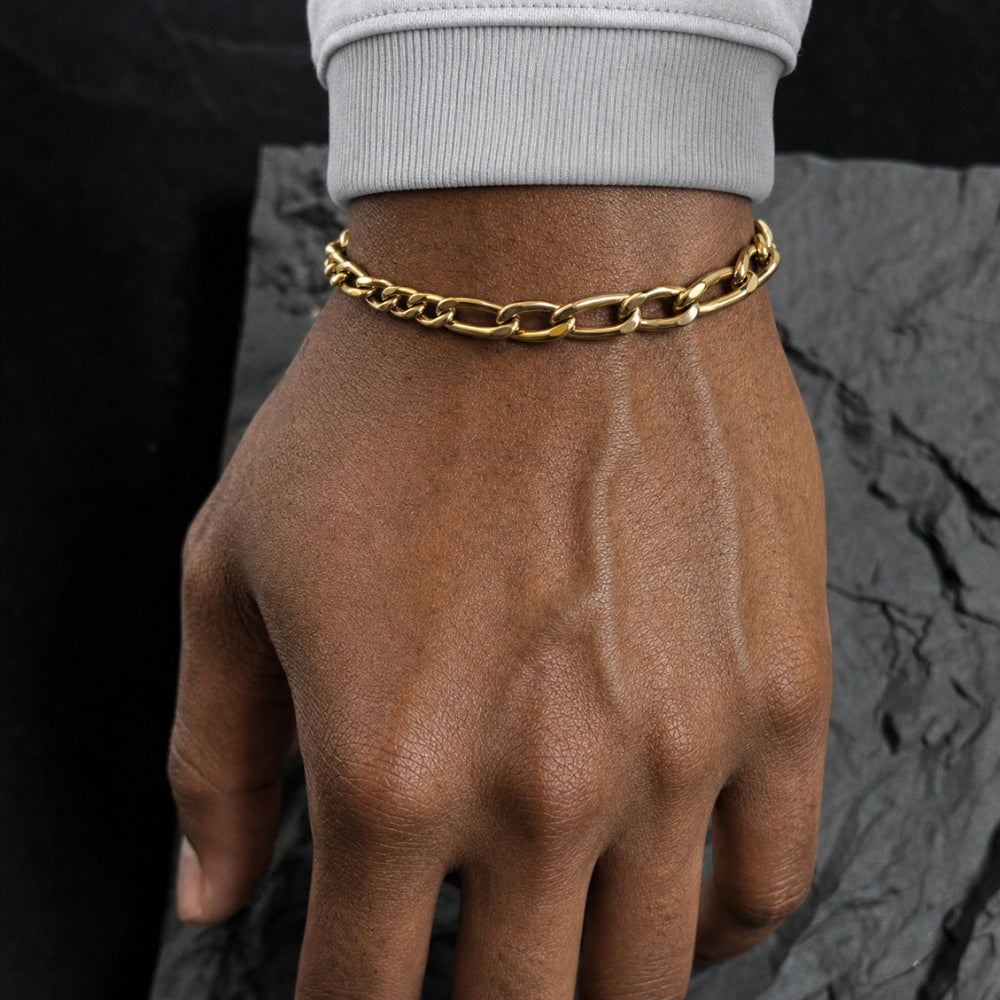 A close-up of a person's hand and wrist wearing a polished gold chain link bracelet, with a light gray ribbed cuff visible at the wrist and a dark textured stone surface in the background.