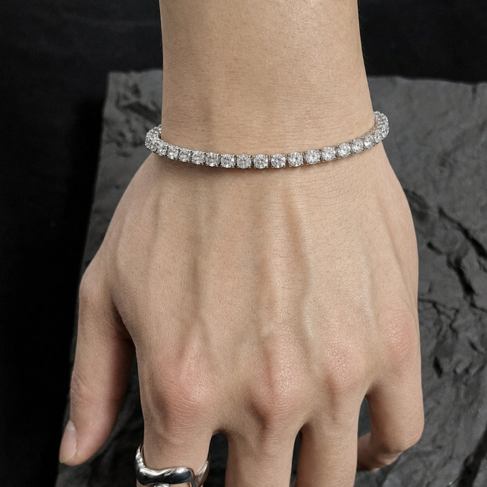 A close-up of a person's hand and wrist wearing a thin silver tennis bracelet with square-cut clear stones, set against a dark textured background.