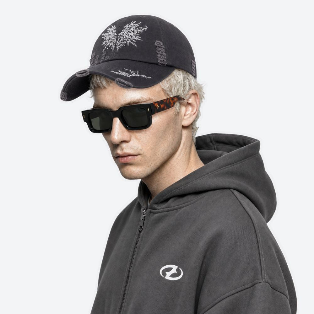 A man wearing a black moth-embroidered baseball cap with sunglasses and a dark hooded sweatshirt