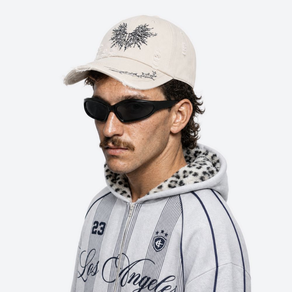 A man wearing a cream moth-embroidered baseball cap with sunglasses and a white varsity jacket