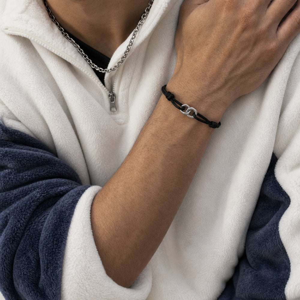 A close-up of a person wearing a black braided cord bracelet with a silver interlocking 'C' clasp, paired with a silver chain necklace and a white and navy fleece pullover.