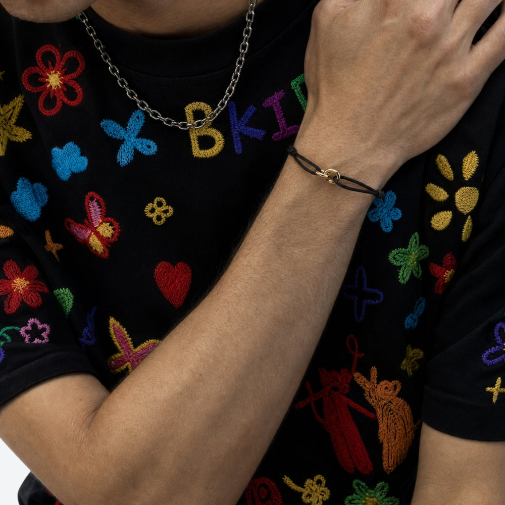 A close-up lifestyle shot of a person wearing a black embroidered t-shirt with colorful floral and letter motifs, a silver chain necklace, and a black cord bracelet with a gold clasp.