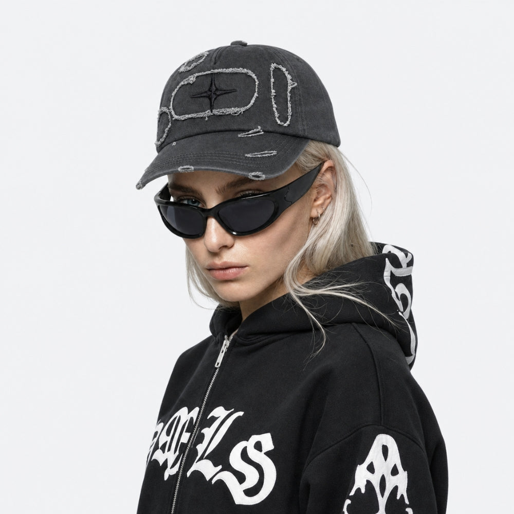 A model wearing a black distressed baseball cap with white embroidered graphic details, black cat-eye sunglasses, and a black hoodie with white graphic lettering, set against a plain light gray background.