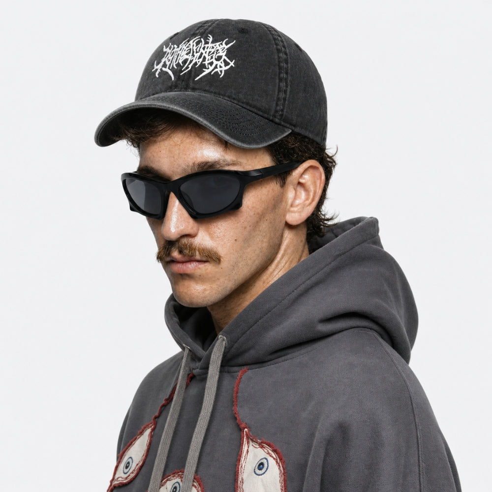 A man wearing a dark gray washed baseball cap with a white graphic logo, black sunglasses, and a dark gray hoodie with a graphic print, shown in a side profile against a light gray background.