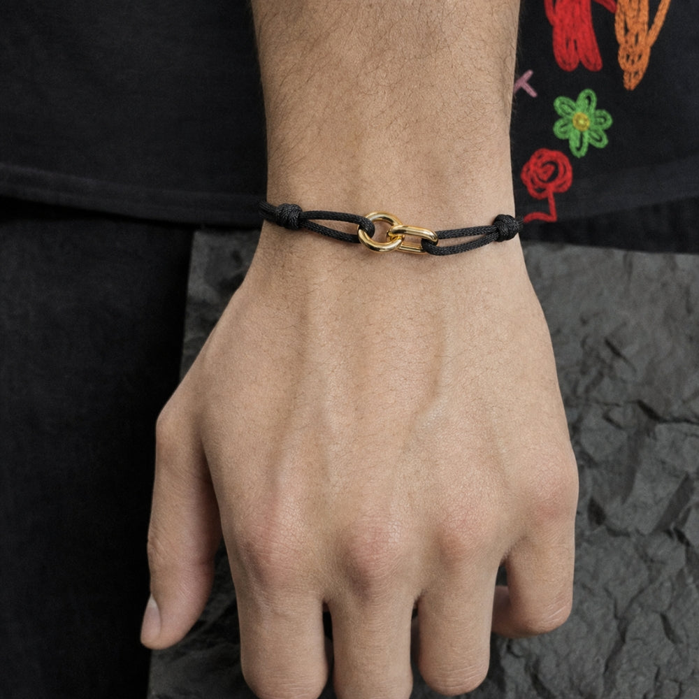 A close-up of a person's wrist wearing a black cord bracelet with a gold-toned interlocking ring clasp, set against a dark shirt and textured gray surface.