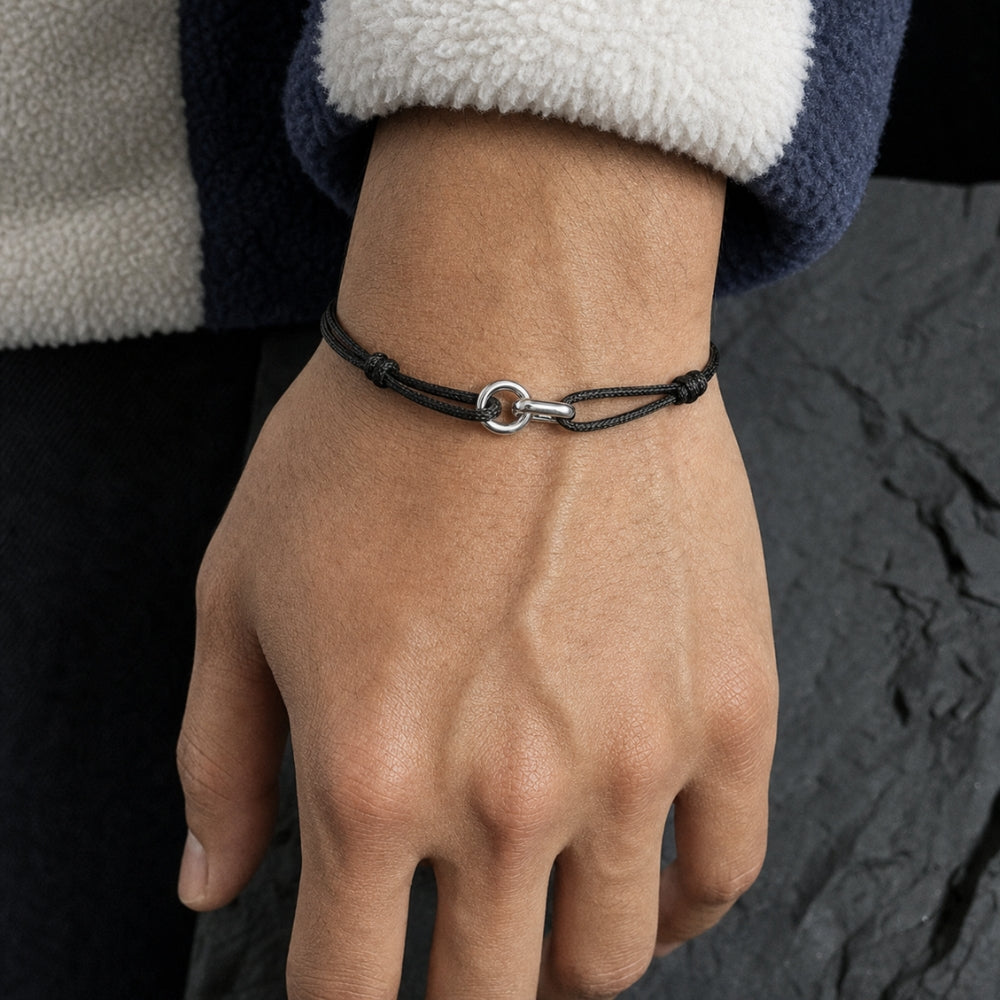 A close-up of a person's hand and wrist wearing a thin black cord bracelet with a silver-toned circular ring clasp, set against a dark textured background.